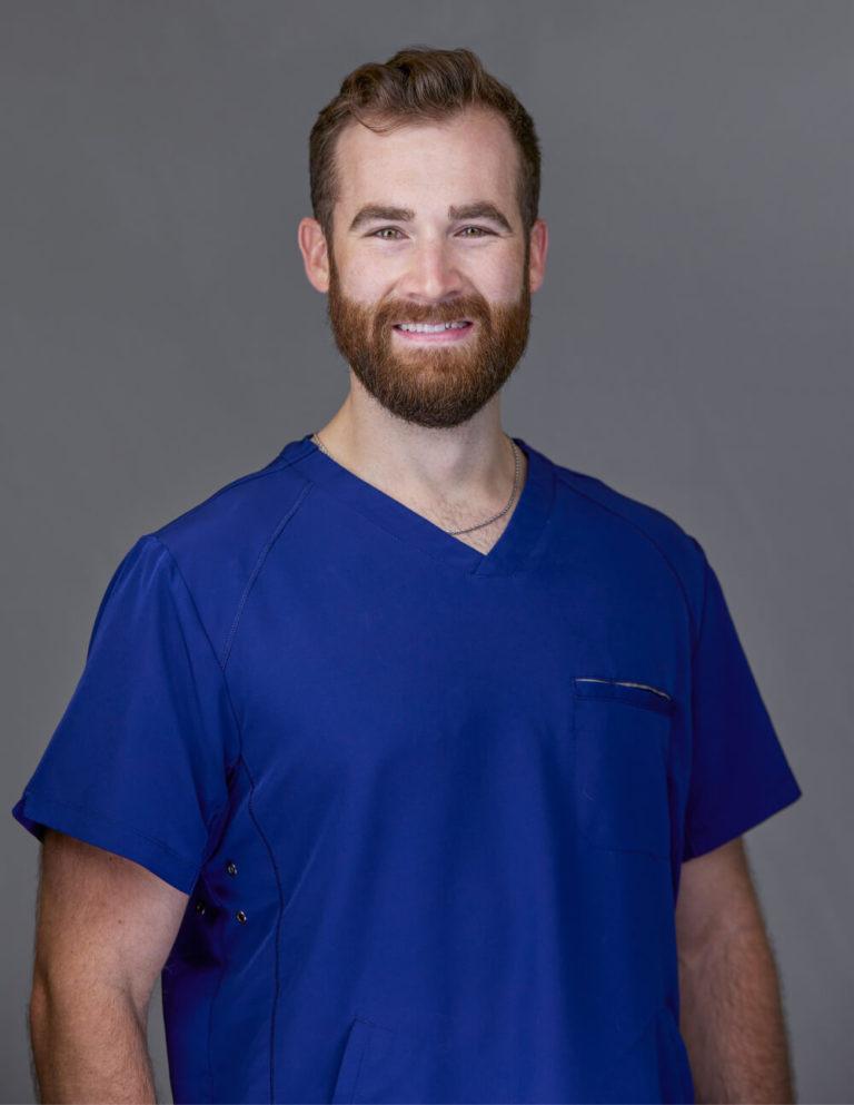 Armored Massage Owner, Stephen Preyer a handsome young man, with brown hair and a beard, dressed in blue scrubs, and shining bright green eyes